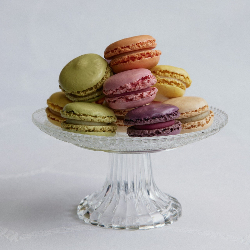 Colorful macarons on a glass cake stand against a light gray background